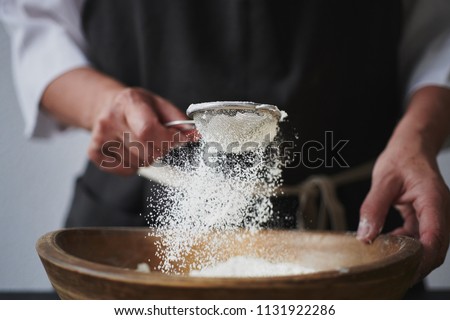 Slow motion shot of aged female hands sifting flour by sieve in wooden bowl. Royalty-Free Stock Photo #1131922286