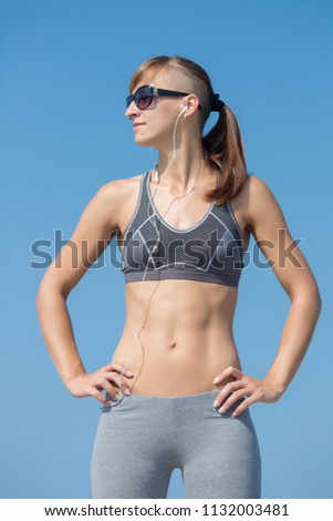 Portrait of sportswoman on background of clear sky. Athletic girl in gray sport bra and trousers posing arms akimbo against sky