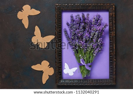 
Bouquet of lavender in wooden frame and paper silhouettes of butterflies. Top view, close-up on dark concrete background.
