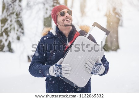 Portrait of young fashionable man on snow holding suitcase. Winter vacation travel concept.