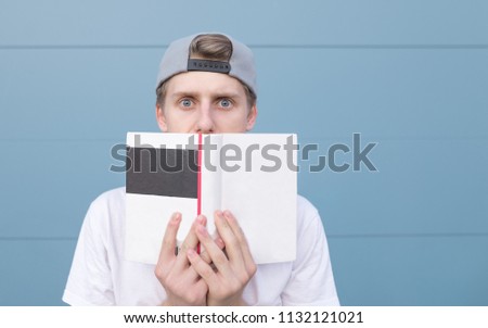 Funny young man with astonished gaze hid behind a book on the background of a blue wall. Portrait of a surprised, funny student with a book on a pastel blue background