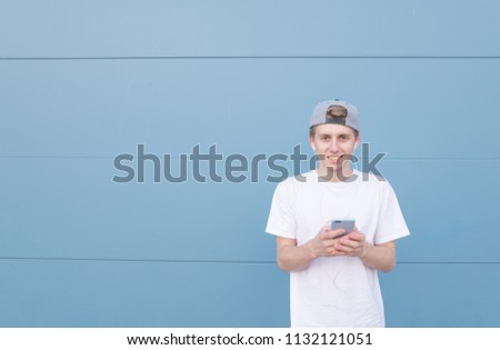 Smiling man stand with a smartphone in his hands on a blue pastel background, listens music in the headphones and looks at the camera.Street portrait of a young man on a background of a blue wall.