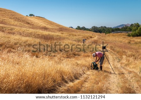 A man is setting up a large format camera on a path in California's golden grasses. Hills rise up behind him. He is wearing a red shirt. A blue sky is in the background.
