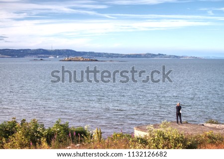 A picture of a lone man taking a photograph of the now deserted island of Inchmickery. The photograph was taken on Cramond Island overlooking the Firth of Forth near Edinburgh, Scotland, UK. 