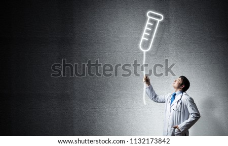 Young confident doctor in white medical uniform interracting with glowing vial symbol whie standing against dark gray wall on background.