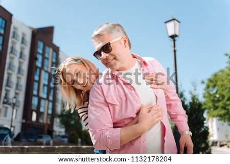 Pleasant emotions. Positive blonde woman smiling to you while hugging her husband