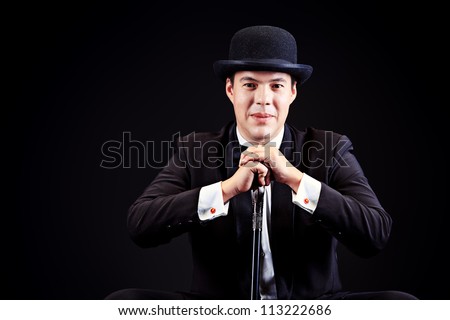 Portrait of an elegant man in black suit and black pot hat holding walking stick. Black background.