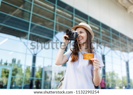 Young smiling traveler tourist woman in hat, take pictures on retro vintage photo camera, hold credit card at international airport. Passenger traveling abroad on weekends getaway. Air flight concept