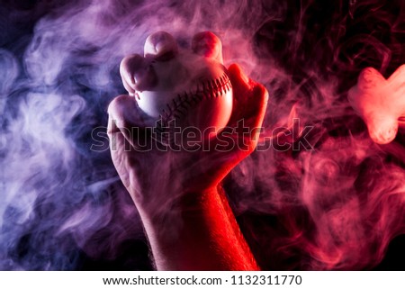 Close-up of a strong male hand holding a white baseball ball  against a background of colored  blue  and red smoke and black isolated background. 