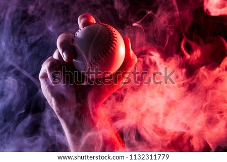 Close-up of a strong male hand holding a white baseball ball  against a background of colored  blue  and red smoke and black isolated background. 
