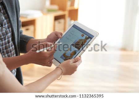 Crop side view of hands of male and female colleagues discussing and editing illustration on tablet screen in hands on blurred backlit background 