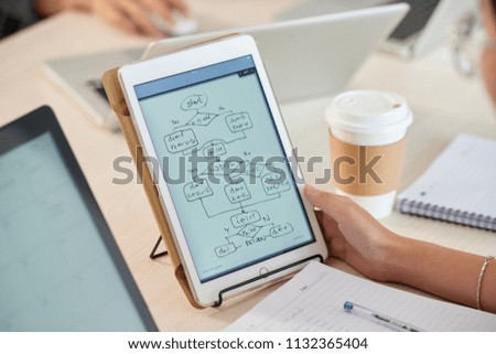 Crop view of female hand holding white tablet with flowchart diagram representing computer program project sitting at desk with laptops on blurred background 