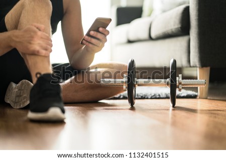 Man using smartphone during workout at home. Online personal trainer or on mobile phone. Internet fitness class or video course. Taking a break. Lazy guy with cellphone while training with dumbbell. Royalty-Free Stock Photo #1132401515