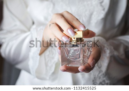 Close up shoot of a luxury perfume flacon being holded in manicured hands of elegant bride wearing white dressing gown Royalty-Free Stock Photo #1132415525