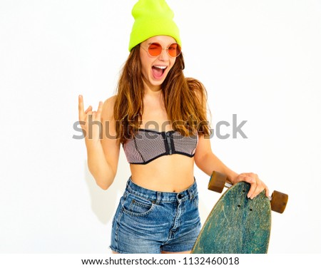 Young stylish smiling girl model in casual summer swimwear clothes and yellow beanie posing with longboard desk, showing rock and roll sign. Isolated on white