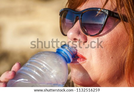 Thirsty young and attractive Caucasian woman in sunglasses drinking cold water from a plastic bottle somewhere in the dessert. Suffering from heat concept image.
