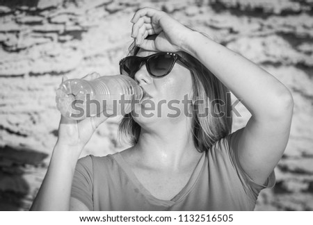 Thirsty young and attractive Caucasian woman in sunglasses drinking cold water from a plastic bottle somewhere in the dessert. Suffering from heat concept image. Black and white photo.