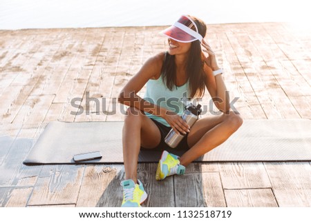 Photo of young sports woman outdoors wearing cap on the beach looking aside drinking water.