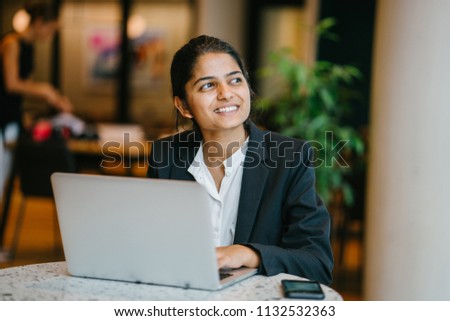 Business portrait of a young Indian Asian woman professional in a suit sitting at a desk in her office. She is replying emails and working on her notebook computer.