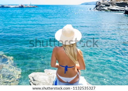 A beautiful blonde tanned girl sitting on the edge of the rock looking at the sea