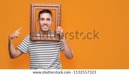 Handsome young man looking through vintage art frame very happy pointing with hand and finger