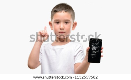 Dark haired little child holding broken smartphone surprised with an idea or question pointing finger with happy face, number one