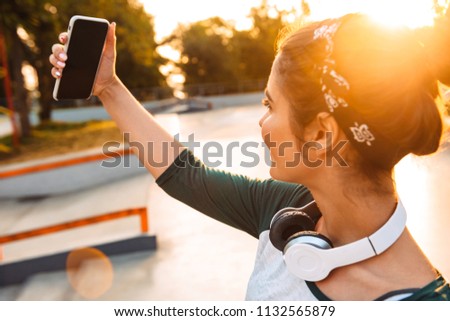 Cheerful young girl with headphones taking a selfie while standing at the park
