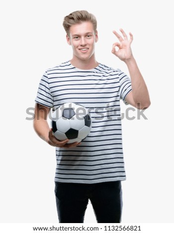 Young handsome blond man holding soccer ball doing ok sign with fingers, excellent symbol
