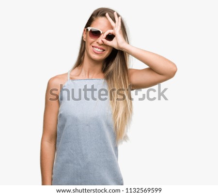 Beautiful young woman wearing sunglasses and denim dress with happy face smiling doing ok sign with hand on eye looking through fingers