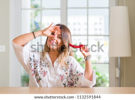 Young woman at home eating red hot chili pepper with happy face smiling doing ok sign with hand on eye looking through fingers