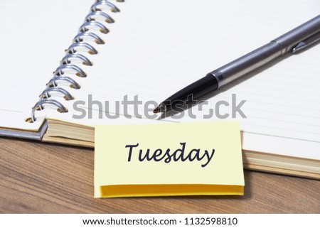 Closeup silver pen on blank page of notebook at wood table with yellow note paper and Tuesday message