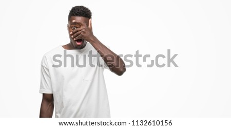 Young african american man wearing white t-shirt peeking in shock covering face and eyes with hand, looking through fingers with embarrassed expression.