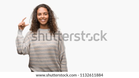 Beautiful young hispanic woman wearing stripes sweater smiling and confident gesturing with hand doing size sign with fingers while looking and the camera. Measure concept.
