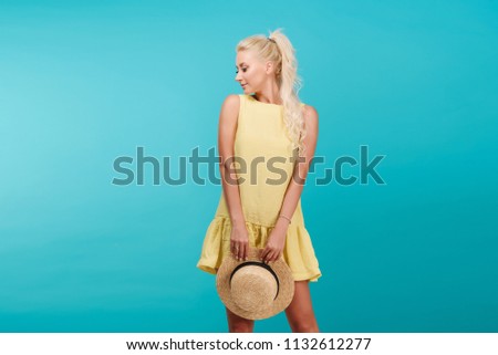 A young slender woman model of a blonde in a summer yellow dress and wearing a hat on a blue background. Model carefree fun