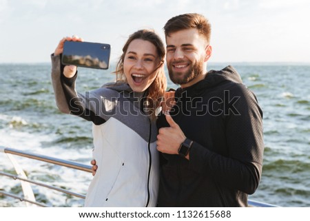 Image of happy young loving couple friends standing outdoors at the beach make selfie by mobile phone showing thumbs up.
