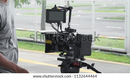 Back angle view of high definition digital video camera which recording or shooting tv commercial production on pan tilt shift professional head tripod at outdoor location and film crew team with set.