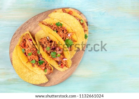 An overhead photo of Mexican tacos with pulled pork, avocado, chili peppers, cilantro, on a teal background with a place for text