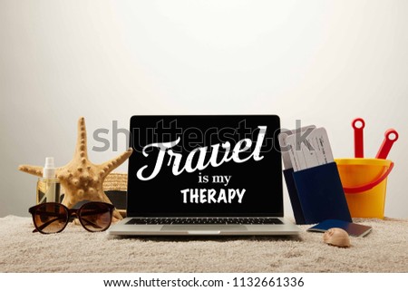 close up view of laptop with "travel is my therapy" lettering, sea star, sunglasses, passports with tickets and toy bucket on sand on grey background