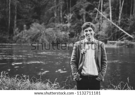 The portrait of attractive groom in a suit and bow tie with boutonniere or buttonhole on jacket, is standing against the background of the forest at nature near river or lake. Black and white photo.