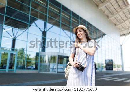 Young smiling traveler tourist woman in hat holding retro vintage photo camera, standing at international airport. Female passenger traveling abroad to travel on weekends getaway. Air flight concept