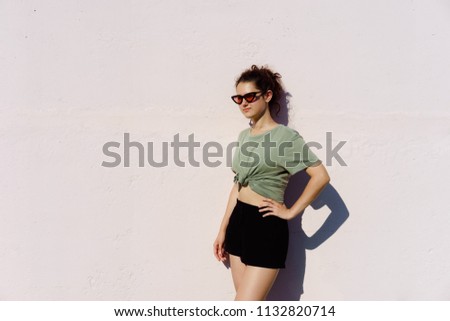 Teen girl in green t-shirt stands on pink wall background in fashionable sunglasses cat's eye, posing in bright Sunny day