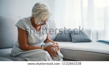 Senior woman holding the knee with pain. Old age, health problem and people concept - senior woman suffering from pain in leg at home. Elderly woman suffering from pain in knee at home Royalty-Free Stock Photo #1132863338