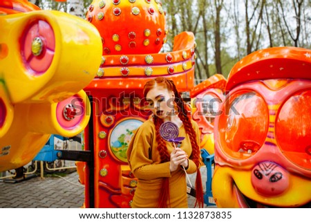 young cheerful red-haired girl posing on the background of the carousel in the amusement Park. Portrait of a woman with a bright make-up and long red hair, who holds a purple candy.