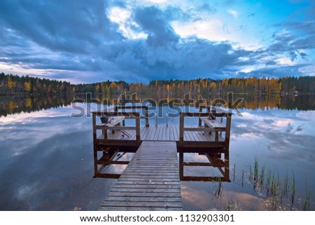 The sunset cloudy autumn sky is reflected in the calm water of the forest lake with a dock and fishing gear in the foreground