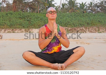 Bald man freak in bright clothes and round glasses at a freak parade, festival, on the beach. India, Goa. An unusual man, the image of a designer's man. The man folded his hands in namaste or prayer.
