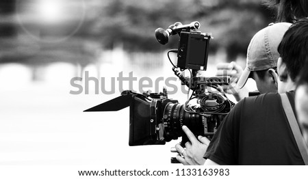 Back angle view of high definition digital video camera which recording or shooting tv commercial production on pan tilt shift professional head tripod at outdoor location and film crew team with set. Royalty-Free Stock Photo #1133163983