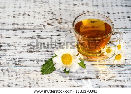 A cup of tea and a saucer, standing on a wooden table, outdoors, with flowers of white daisies, in the rays of sunlight.