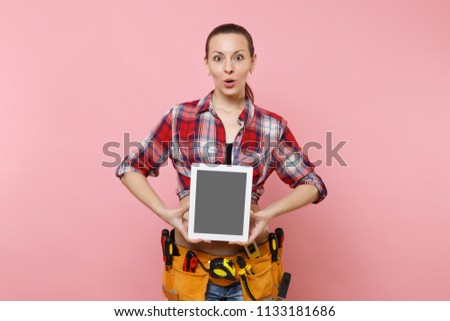 Woman with kit tools belt full of variety instruments hold tablet pc computer with blank black empty screen display touchscreen isolated on pink background. Female doing male work. Renovation concept