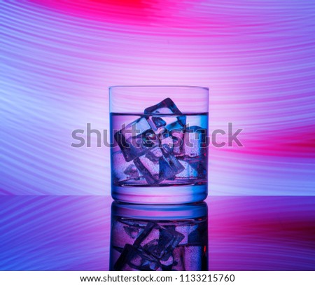 A glass with a drink and ice cubes on a red background with blue lights.