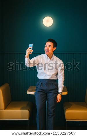 A professional Chinese Asian man in a shirt and pants takes a selfie in the office in a trendy booth. 